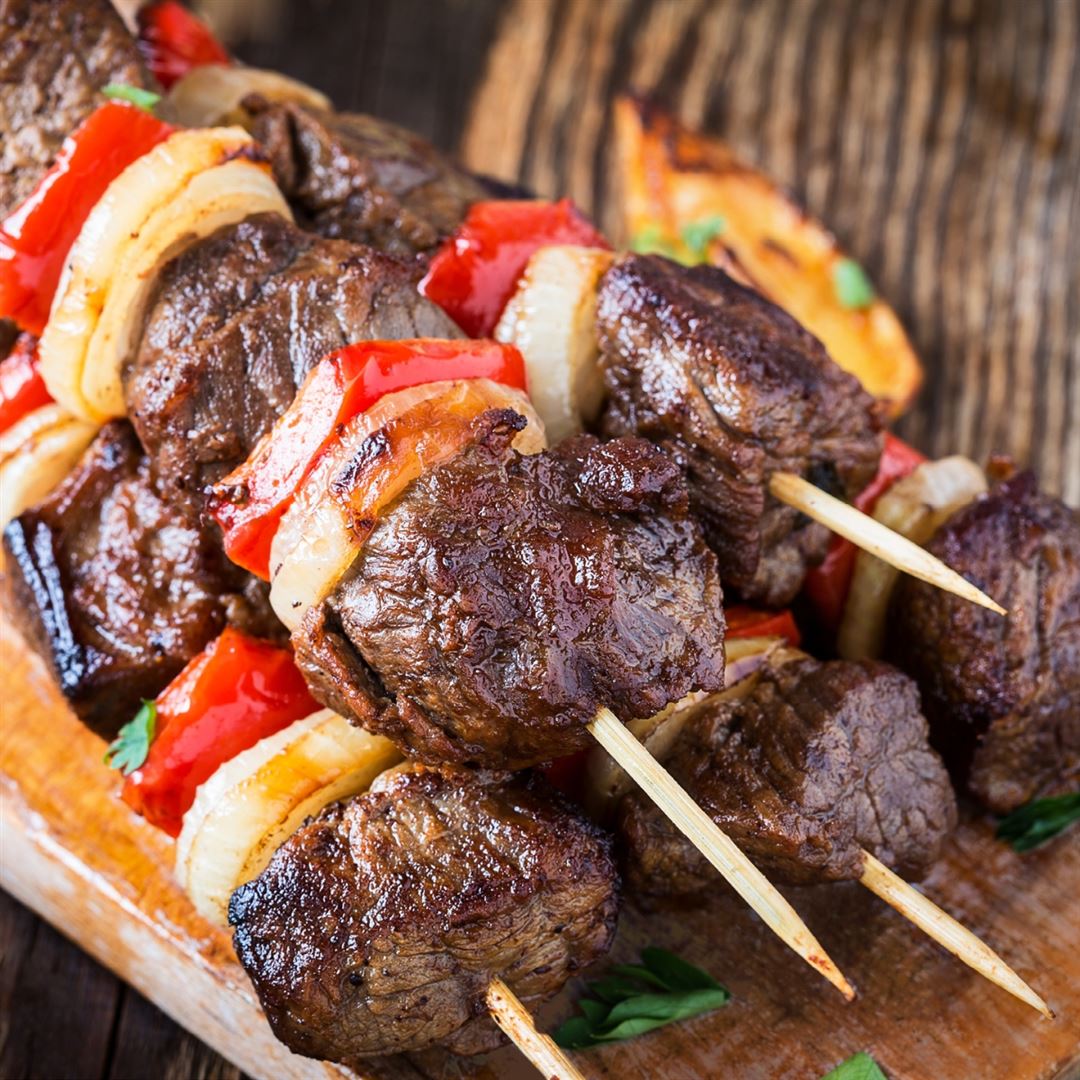 Close-up of grilled beef kebabs on wooden skewers, featuring chunks of meat, red bell peppers, and onion slices, served on a rustic wooden board. The food looks juicy and freshly cooked.