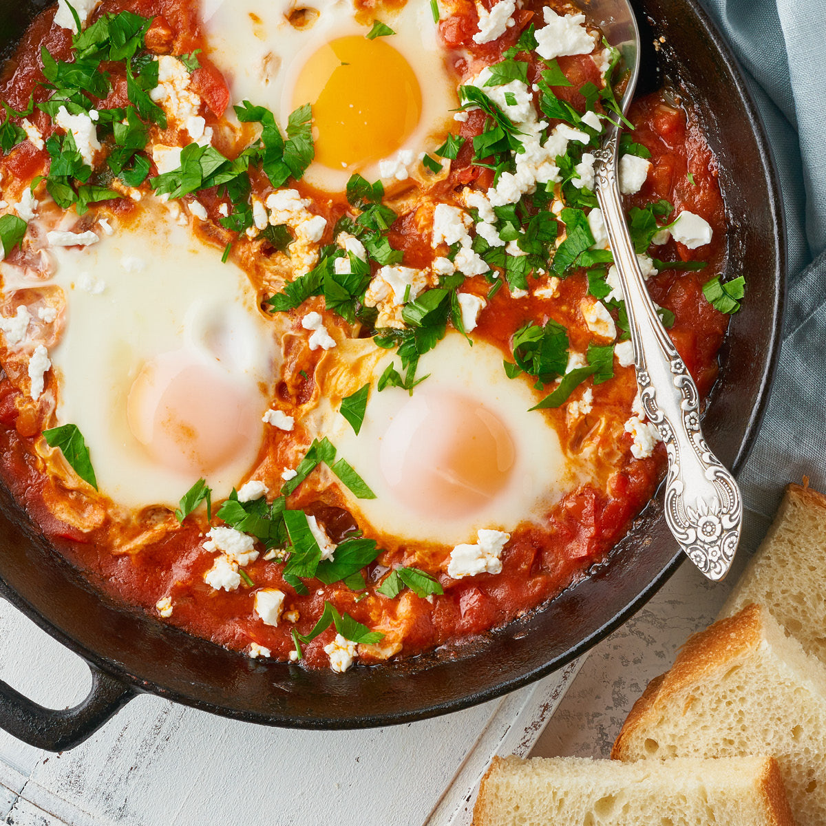 A skillet of shakshuka with three poached eggs in a tomato sauce, topped with chopped parsley and crumbled feta cheese, served alongside slices of bread.