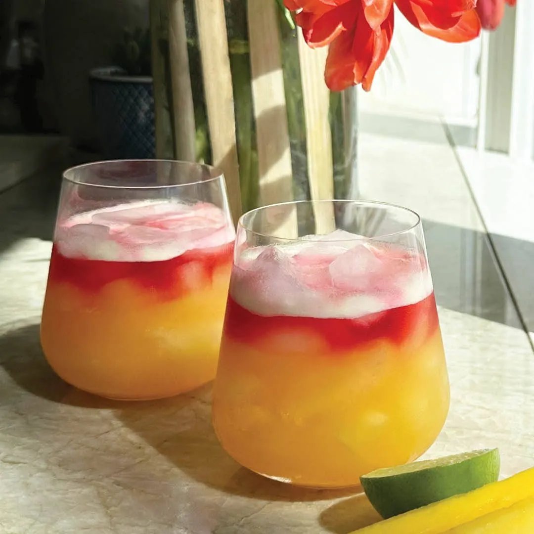 Two glasses of a colorful layered drink with ice, featuring yellow-orange juice at the bottom and a red layer on top, sit on a sunlit countertop. Slices of lemon and lime are nearby, with orange flowers in the background.