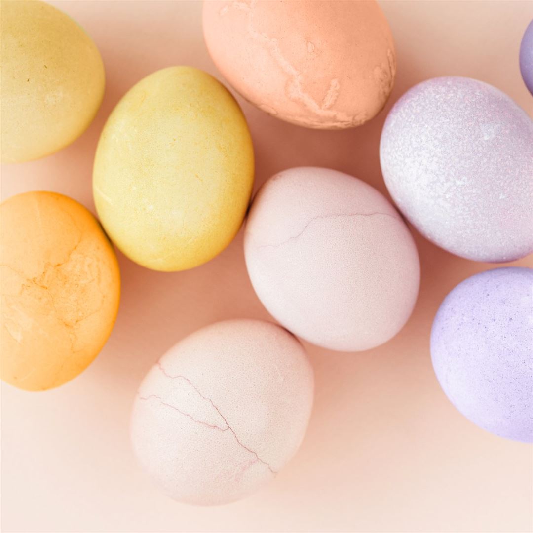 A group of pastel-colored eggs, including shades of yellow, orange, pink, and purple, arranged on a light peach background.