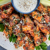 A plate of crispy chicken wings garnished with chopped herbs and grated cheese, served with a small bowl of creamy dipping sauce and celery sticks on the side.