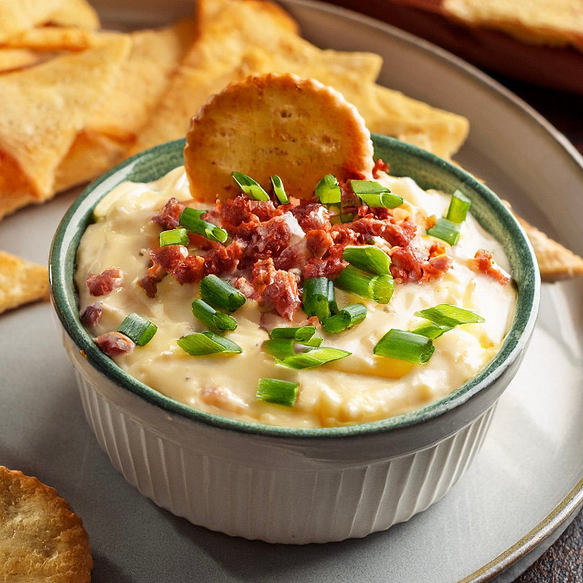 A bowl of creamy dip topped with chopped bacon and green onions, garnished with a round cracker. Tortilla chips are arranged on a plate around the bowl.