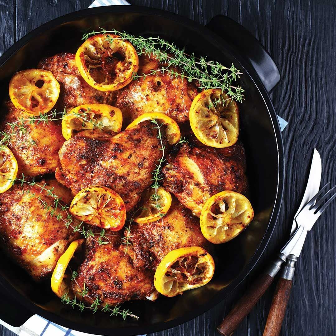 A black roasting pan filled with seasoned baked chicken thighs, garnished with lemon slices and fresh thyme sprigs, sits on a dark wooden table beside a fork and knife with wooden handles.