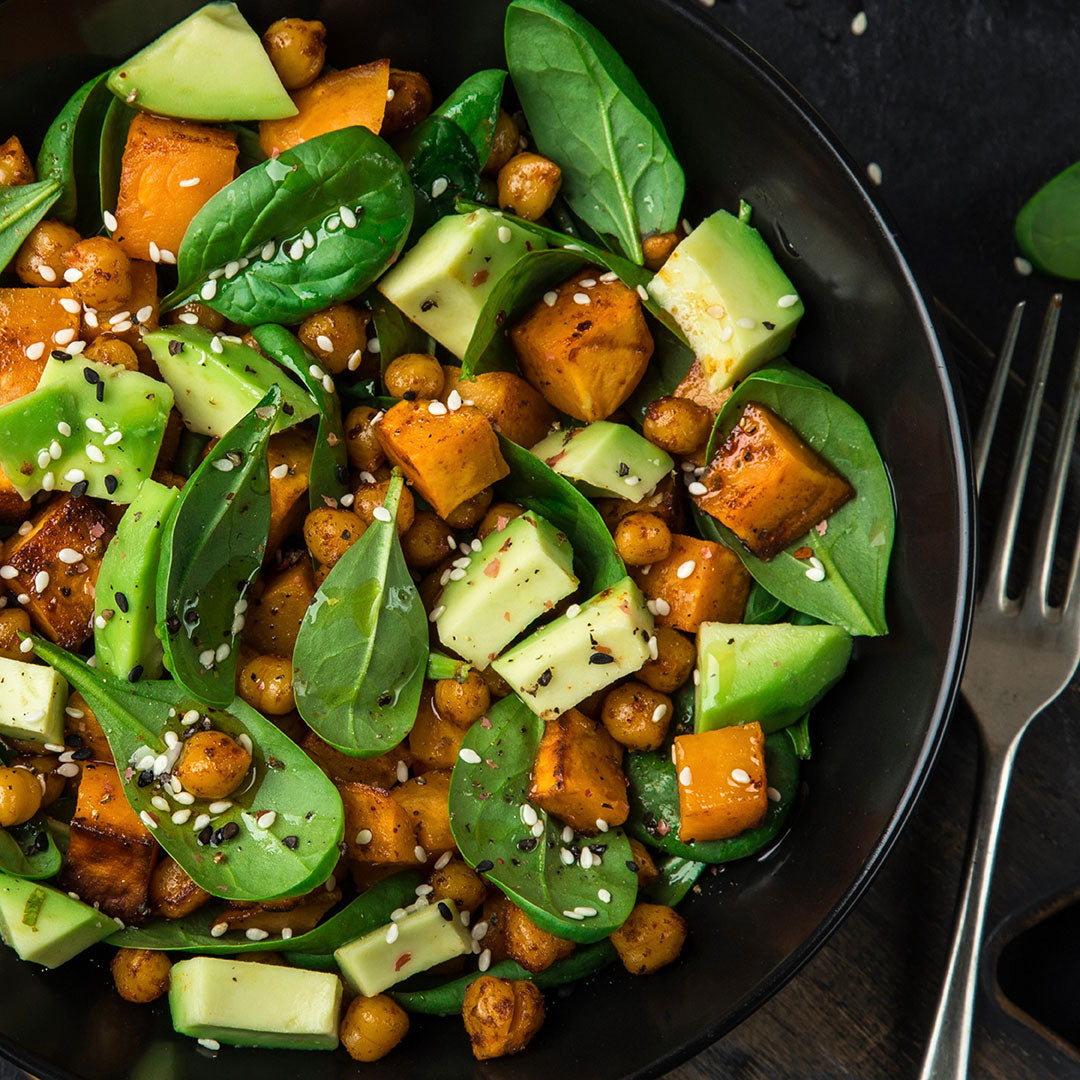 A bowl filled with fresh spinach leaves, roasted sweet potato cubes, chickpeas, avocado chunks, and a sprinkle of sesame seeds, with a fork beside the bowl on a dark surface.