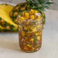 A glass jar filled with pineapple salsa, containing diced pineapple, red onion, and herbs, sits on a light surface. A fresh pineapple, partially sliced, is in the background.