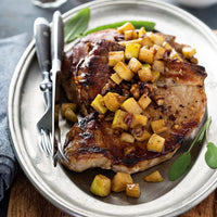 Apple Baked Pork Chops (Kit): A juicy pork chop seasoned with autumn spices, topped with diced apples and pecans, served on a silver plate with fresh sage. Fork and knife provided alongside.