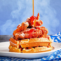 Hot 'N Spicy Chicken and Waffles (Kit) features crispy fried chicken atop waffles, drizzled with spicy maple syrup and served on a white plate against a blue checkered cloth and smoky blue background.