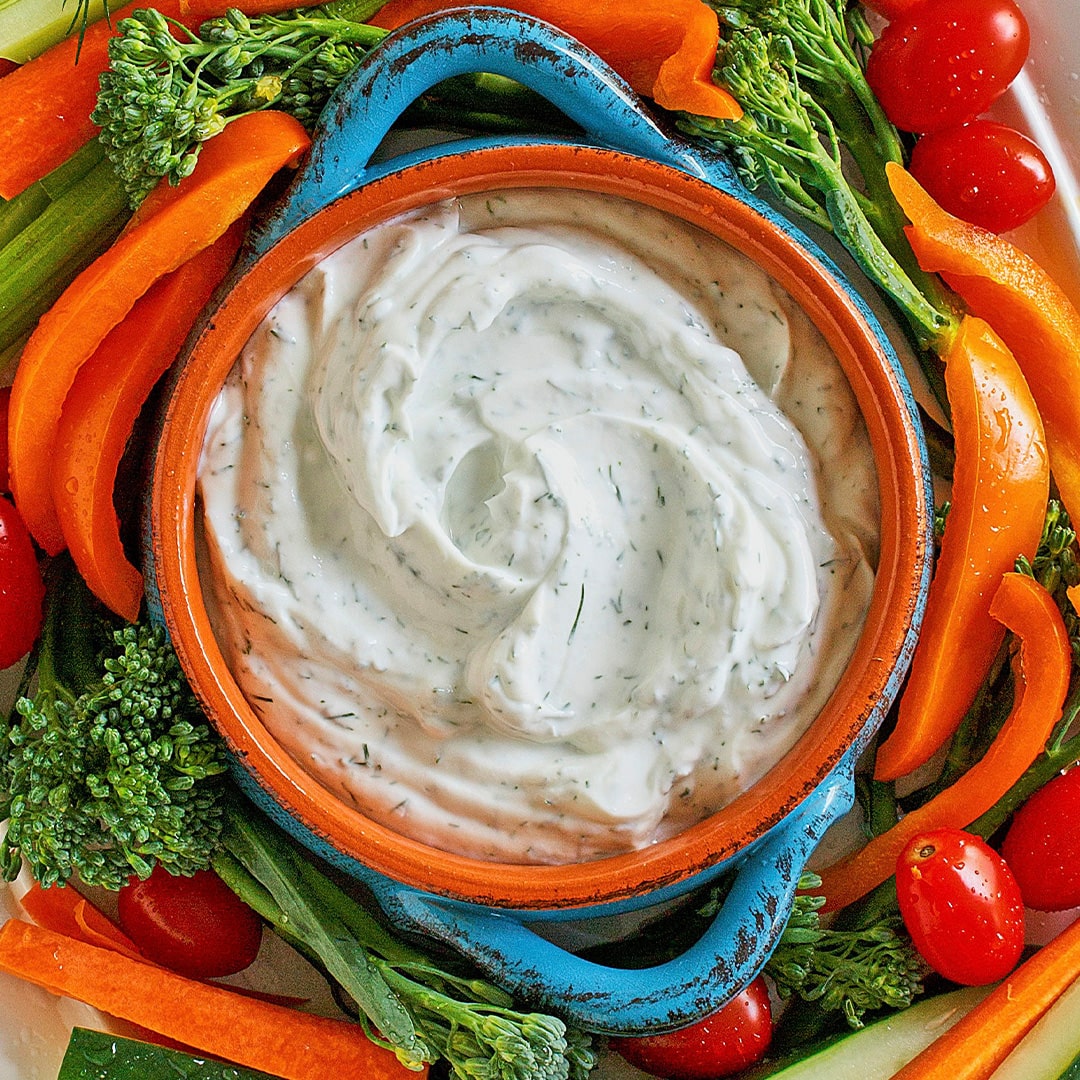 A colorful platter with a bowl of creamy ranch dip surrounded by fresh vegetables, including orange bell pepper slices, cherry tomatoes, broccolini, carrot sticks, and celery.