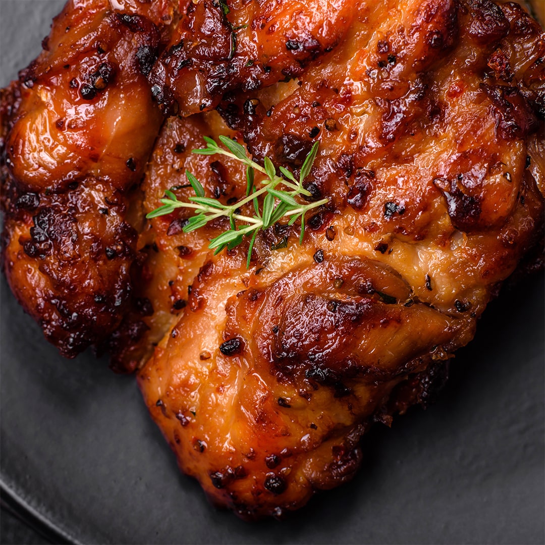 Close-up of a piece of grilled, glazed chicken garnished with a small sprig of fresh thyme, served on a dark plate. The chicken is golden-brown with a slightly charred, caramelized surface.