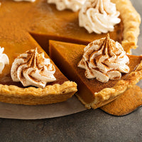 A close-up of No Bake Pumpkin Pie (Kit) with a slice served, topped with whipped cream and Pumpkin Pie Spice Blend on a golden, flaky crust—an ideal Thanksgiving dessert on a dark surface.