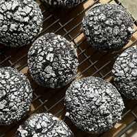 Black Cocoa Crinkle Cookies (Kit) with a cracked surface and powdered sugar are cooling on a wire rack in natural sunlight.