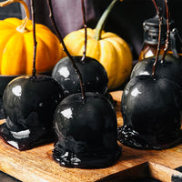 Glossy black candy apples on sticks are arranged on a wooden board, with two small pumpkins and a dark bottle in the background, creating a Halloween-themed scene enhanced by the Black Cocoa Caramel Sauce (Kit).