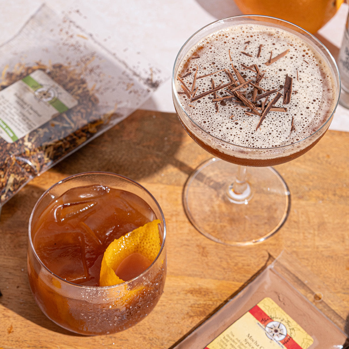 Two cocktails on a wooden surface: one in a coupe glass topped with chocolate shavings, the other in a rocks glass with ice and an orange twist. Nearby are packets of loose leaf tea and tea packaging.