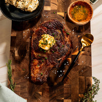 Cowboy Butter Steak (Kit): Grilled steak topped with herb-infused cowboy butter and signature steak seasoning, served on a wooden board and finished with sea salt and fresh chives. Accompanied by a gold spoon, a small bowl of sauce, and a rosemary sprig for garnish.