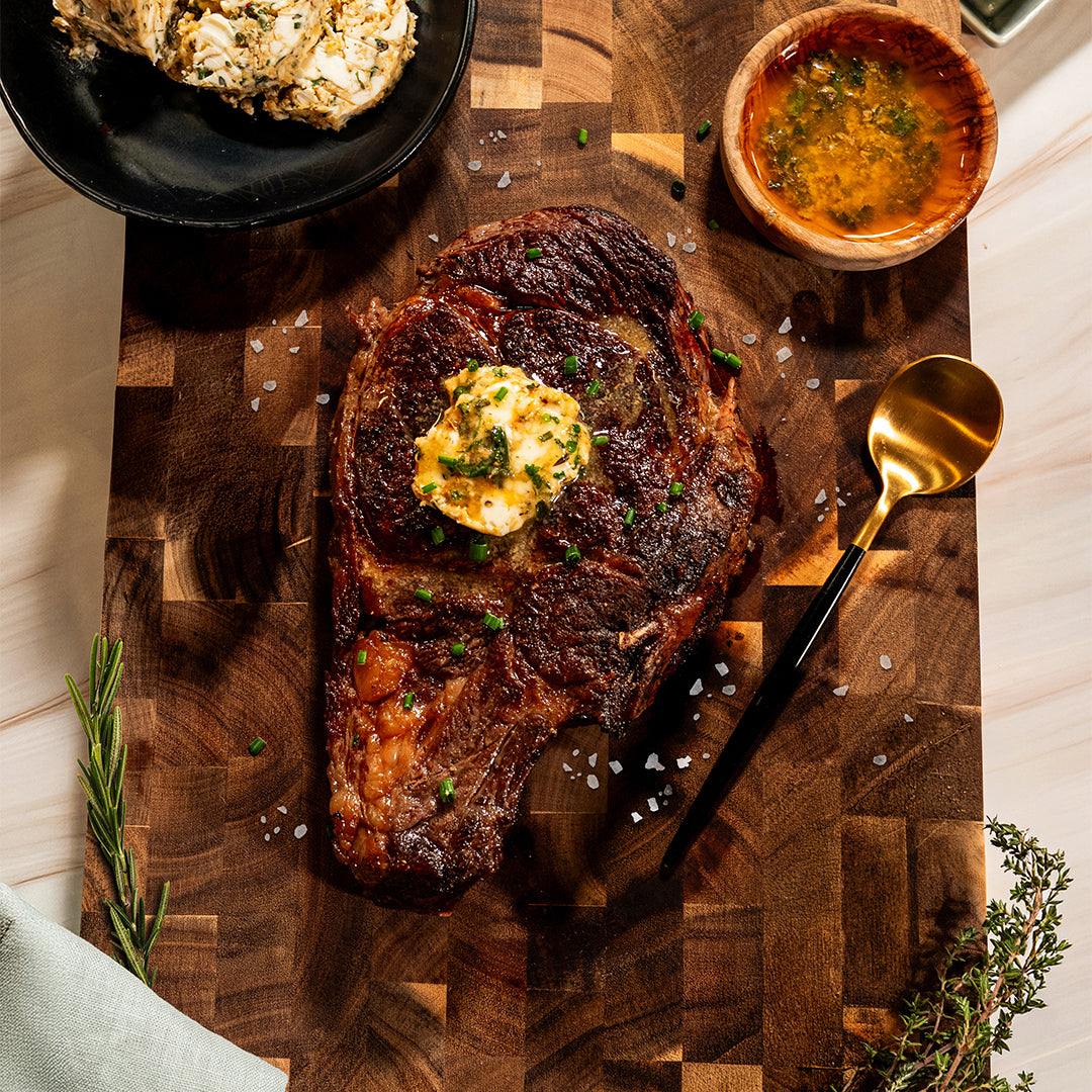Cowboy Butter Steak (Kit): Grilled steak topped with herb-infused cowboy butter and signature steak seasoning, served on a wooden board and finished with sea salt and fresh chives. Accompanied by a gold spoon, a small bowl of sauce, and a rosemary sprig for garnish.