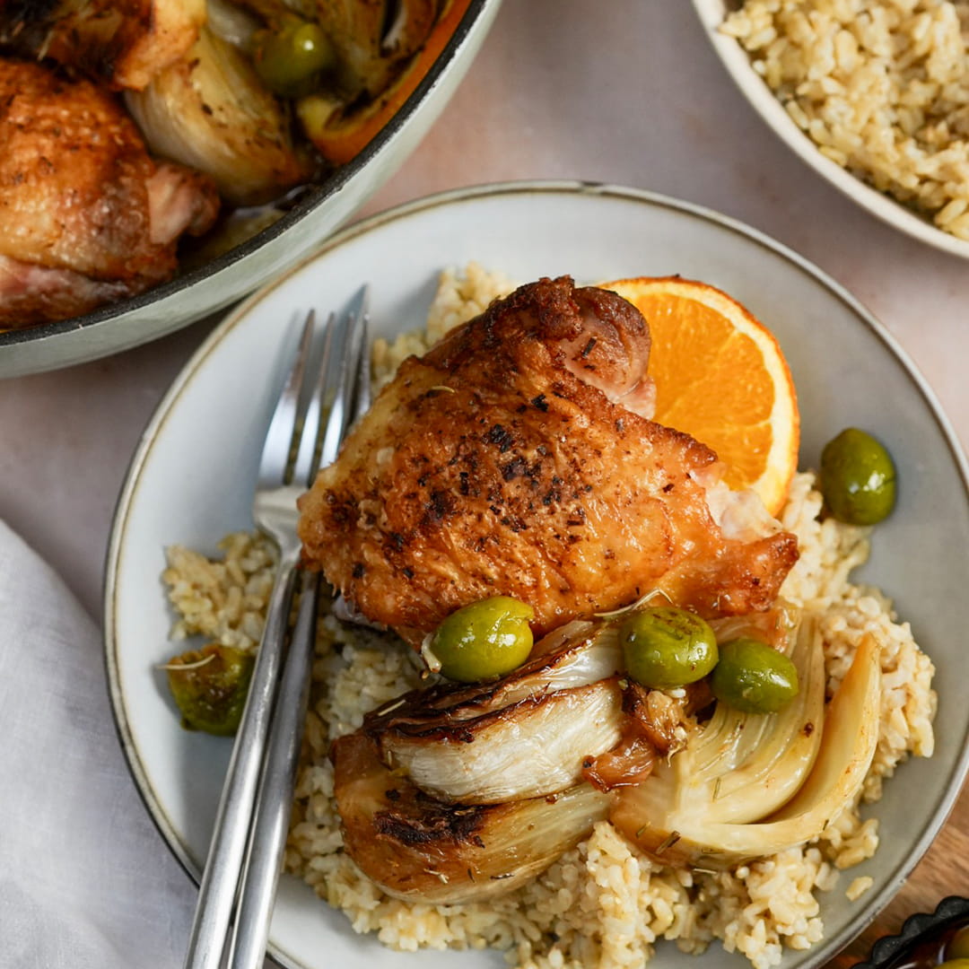 A plated prepared meal featuring a roasted chicken thigh on rice with fennel, olives, and an orange slice, with a pan of food and another plate blurred in the background.