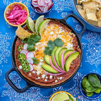 Southwest Cheesy Bean Dip on a Blue Background with bows of Chips Topped with Avacado, Cilantro Leaves and Radishes.