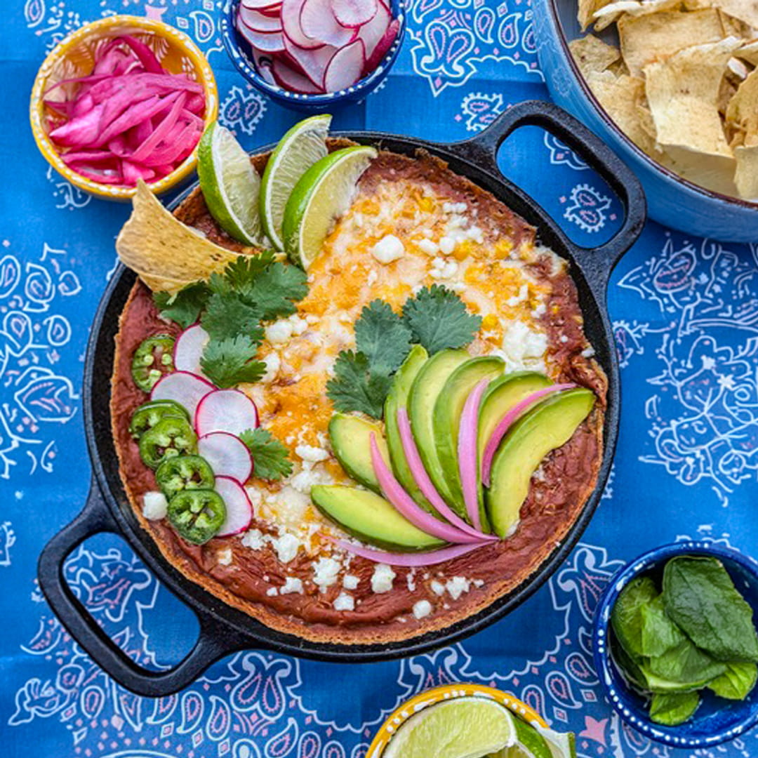 Southwest Cheesy Bean Dip on a Blue Background with bows of Chips Topped with Avacado, Cilantro Leaves and Radishes.