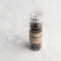 A clear bottle labeled "4 Peppercorn Spice Blend" stands upright on a light marble kitchen surface, filled with peppercorns. Its transparent grinder lid rests nearby.