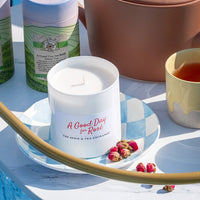 Candle labeled 'A Good Day for Rosé' on a decorative plate with tea and tea packaging in the background.