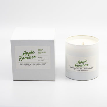 A candle in a white glass jar next to its white packaging box, both featuring the 'Apple Rancher' brand logo in green script on a clean white background.