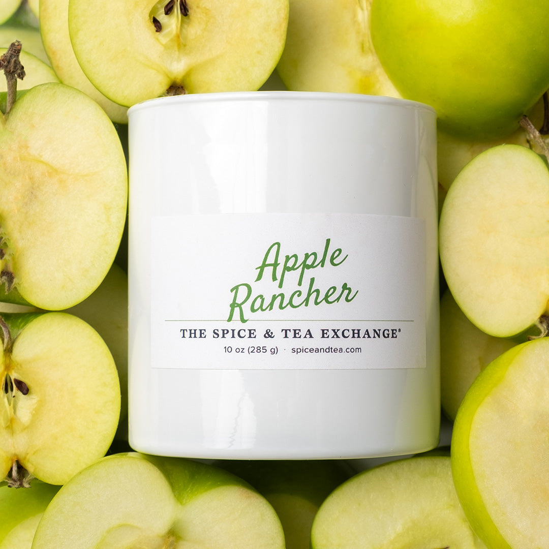 A white candle jar labeled 'Apple Rancher' by The Spice & Tea Exchange, surrounded by fresh green apples and apple slices.