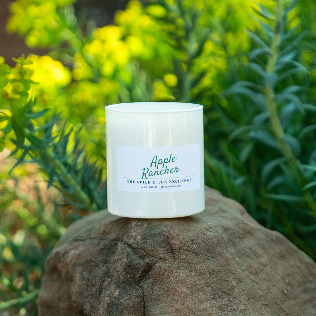 A white glass jar candle labeled 'Apple Rancher' by The Spice & Tea Exchange, resting on a large brown rock with a blurred background of green and yellow foliage.