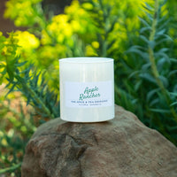 A white glass jar candle labeled 'Apple Rancher' by The Spice & Tea Exchange, resting on a large brown rock with a blurred background of green and yellow foliage.