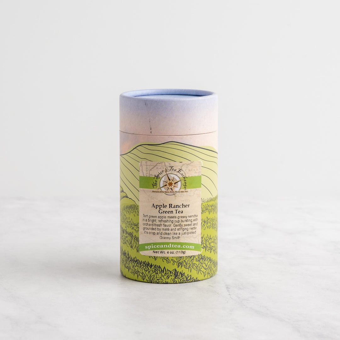 A cylindrical cardboard tin of Apple Rancher Green Tea on a light marble surface against a white background. The tin features a scenic green hill illustration and a label with a compass rose logo.
