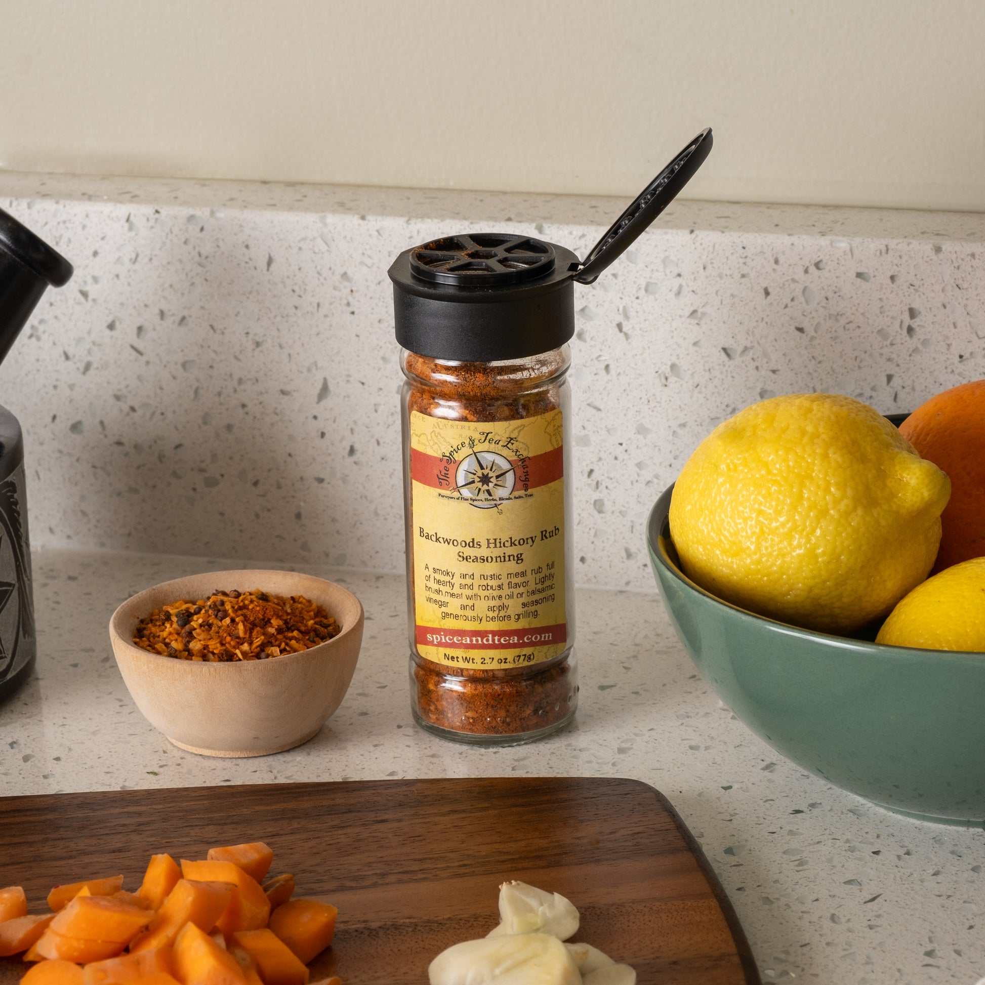 A jar of Backwoods Hickory Rub Seasoning sits on a kitchen counter beside lemons, oranges, a wooden bowl of spices, chopped carrots, and garlic cloves.