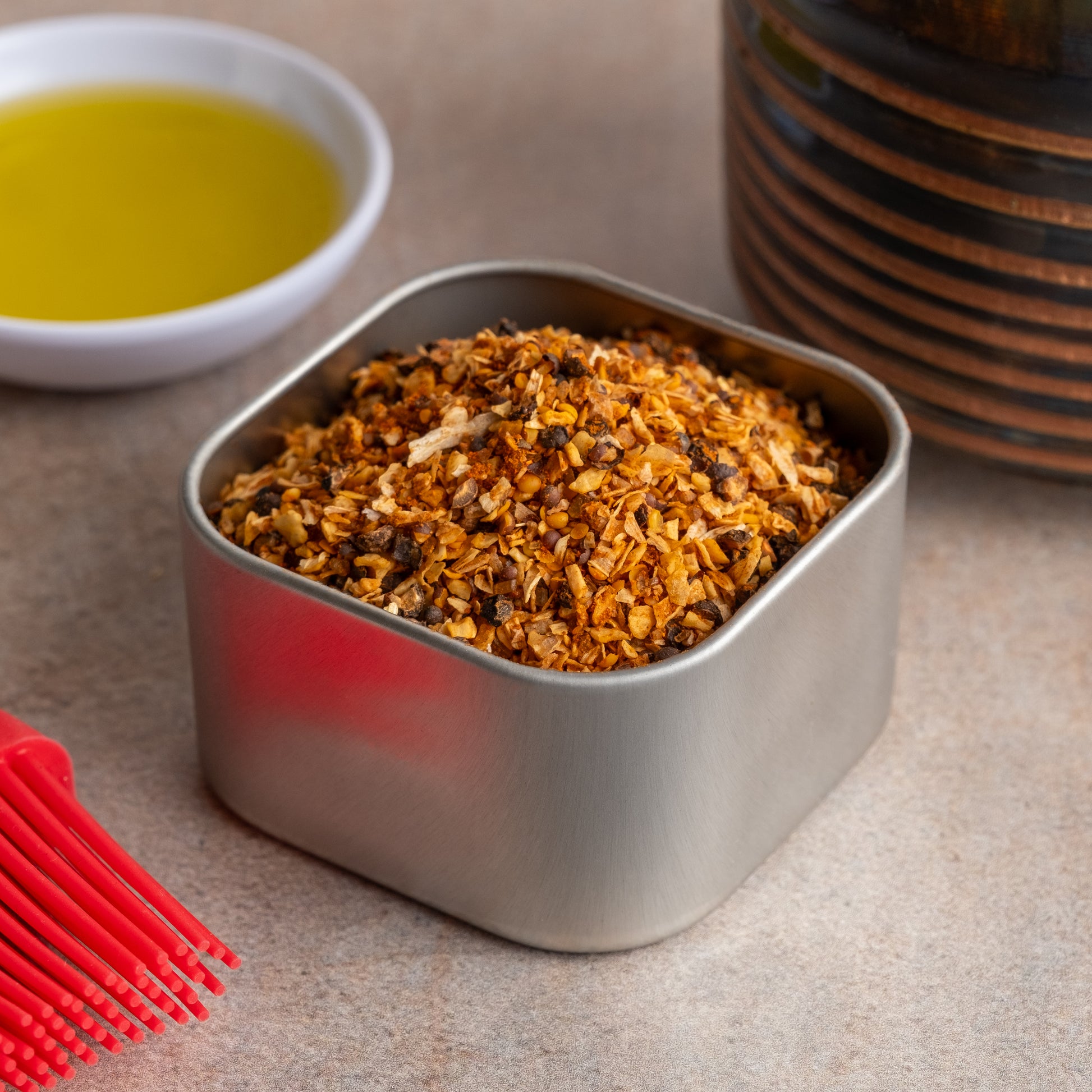 A metal container of Backwoods Hickory Rub Seasoning, filled with coarse orange-brown mix, sits on the countertop next to a bowl of olive oil, a red basting brush, and a striped ceramic jar.