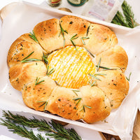 A baked bread roll arranged in a ring around a round cheese center, topped with rosemary sprigs and herbs and seasoning, set in a white baking dish with parchment paper.