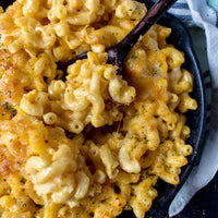 Close-up of creamy macaroni pasta in a black skillet with melted cheese, herbs, and a dark wooden spoon.
