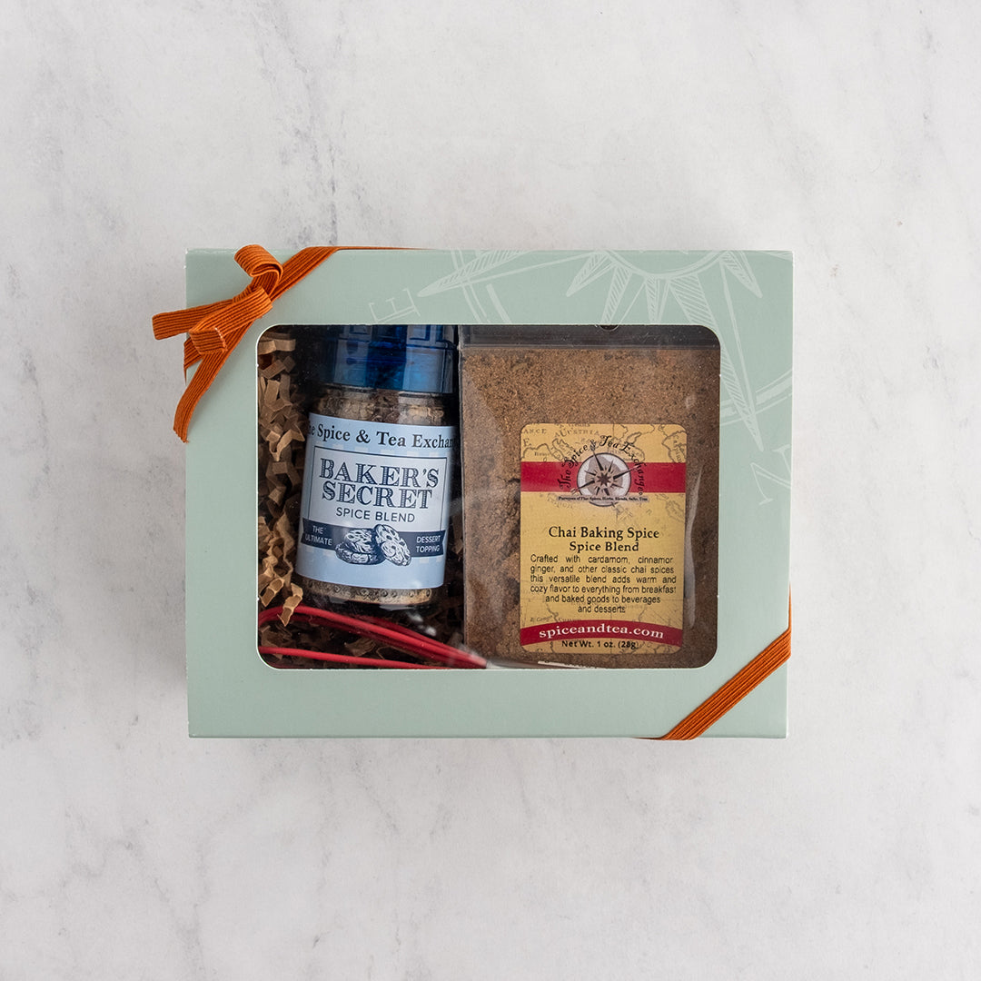 A light green gift box containing a bottle of The Spice & Tea Bakers Secret Spice Blend and a bag of Chai Baking Spice Blend, tied with an orange ribbon on a white marble background.