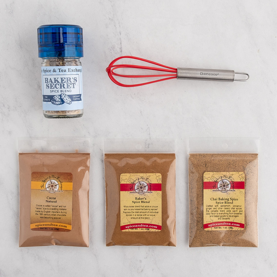 A baking spice kit laid out on a white marble surface, including a small glass bottle of Baker's Secret Spice Blend with a blue lid, a red mini whisk, and three clear bags of spices: Cocoa Natural, Baker's Spice Blend, and Chai Baking Spice Spice Blend.