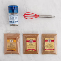 A baking spice kit laid out on a white marble surface, including a small glass bottle of Baker's Secret Spice Blend with a blue lid, a red mini whisk, and three clear bags of spices: Cocoa Natural, Baker's Spice Blend, and Chai Baking Spice Spice Blend.