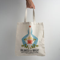 A hand holds the Balanced & Bright Tote Bag from The Spice & Tea Exchange, featuring blue hands holding an orange cup, plants, a burst of light, and the words “LESS STRESS. MORE FLAVOR. SPICEMADETEA.COM.”.