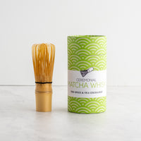 A bamboo matcha whisk stands next to its green and white patterned cylindrical packaging box on a light marble surface against a white wall. The box features a white label with the text 'Ceremonial Matcha Whisk' and 'The Spice & Tea Exchange'.