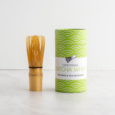 A bamboo matcha whisk stands next to its green and white patterned cylindrical packaging box on a light marble surface against a white wall. The box features a white label with the text 'Ceremonial Matcha Whisk' and 'The Spice & Tea Exchange'.
