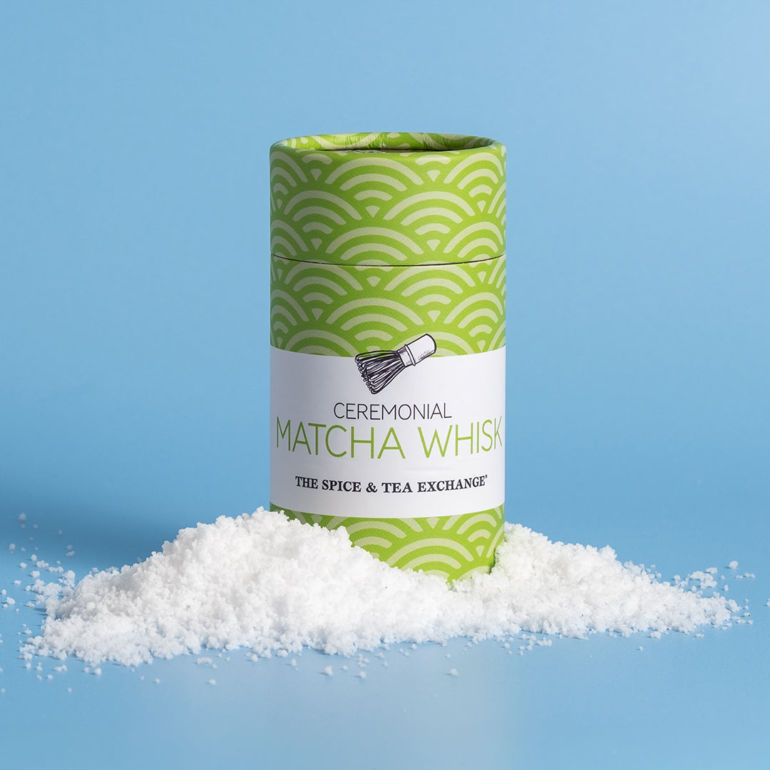 A cylindrical tin of ceremonial matcha whisk by The Spice & Tea Exchange, standing upright on a pile of white matcha powder against a light blue background.