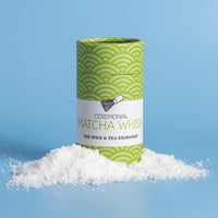 A cylindrical tin of ceremonial matcha whisk by The Spice & Tea Exchange, standing upright on a pile of white matcha powder against a light blue background.