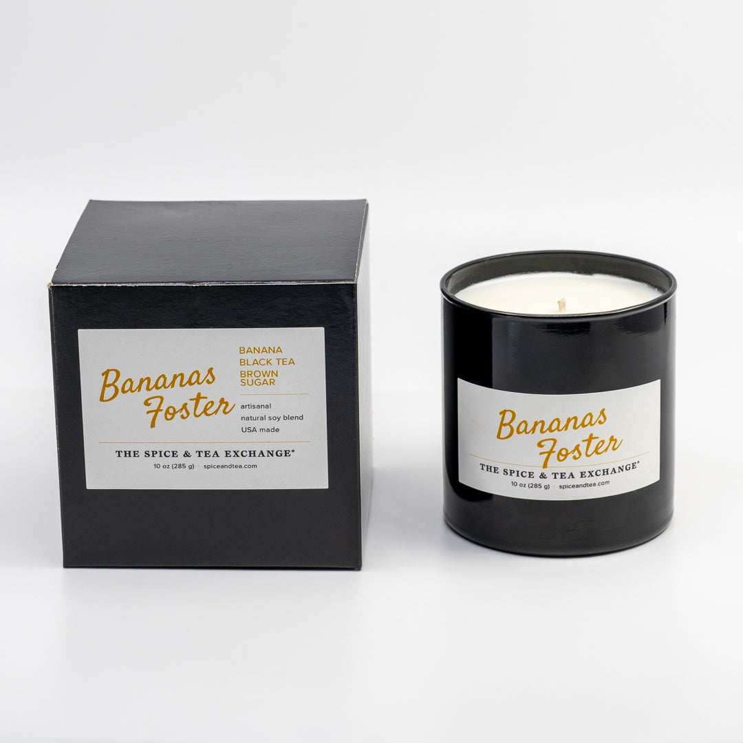 A black glass jar candle with a white label next to its black box packaging, both featuring the Banana Foster scent by The Spice & Tea Exchange.