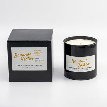 A black glass jar candle with a white label next to its black box packaging, both featuring the Banana Foster scent by The Spice & Tea Exchange.