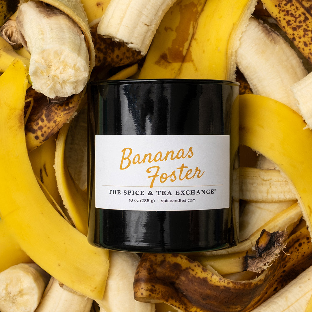 A black candle jar labeled 'Bananas Foster' by The Spice & Tea Exchange, surrounded by fresh banana peels and sliced bananas in a styled flat-lay composition.