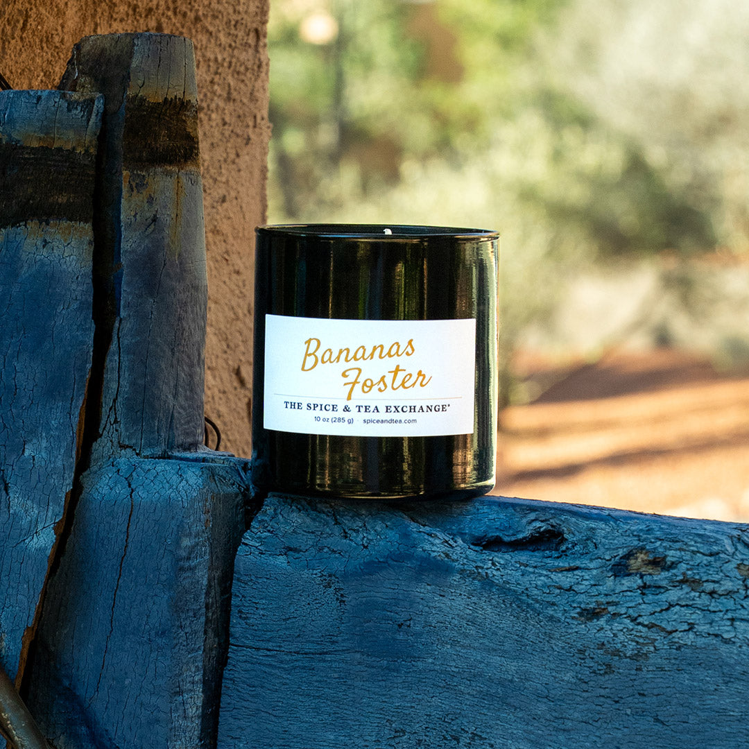 A black glass jar candle with a white label reading 'Bananas Foster' and 'The Spice & Tea Exchange' sits on a weathered blue wooden ledge outdoors.