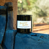 A black glass jar candle with a white label reading 'Bananas Foster' and 'The Spice & Tea Exchange' sits on a weathered blue wooden ledge outdoors.