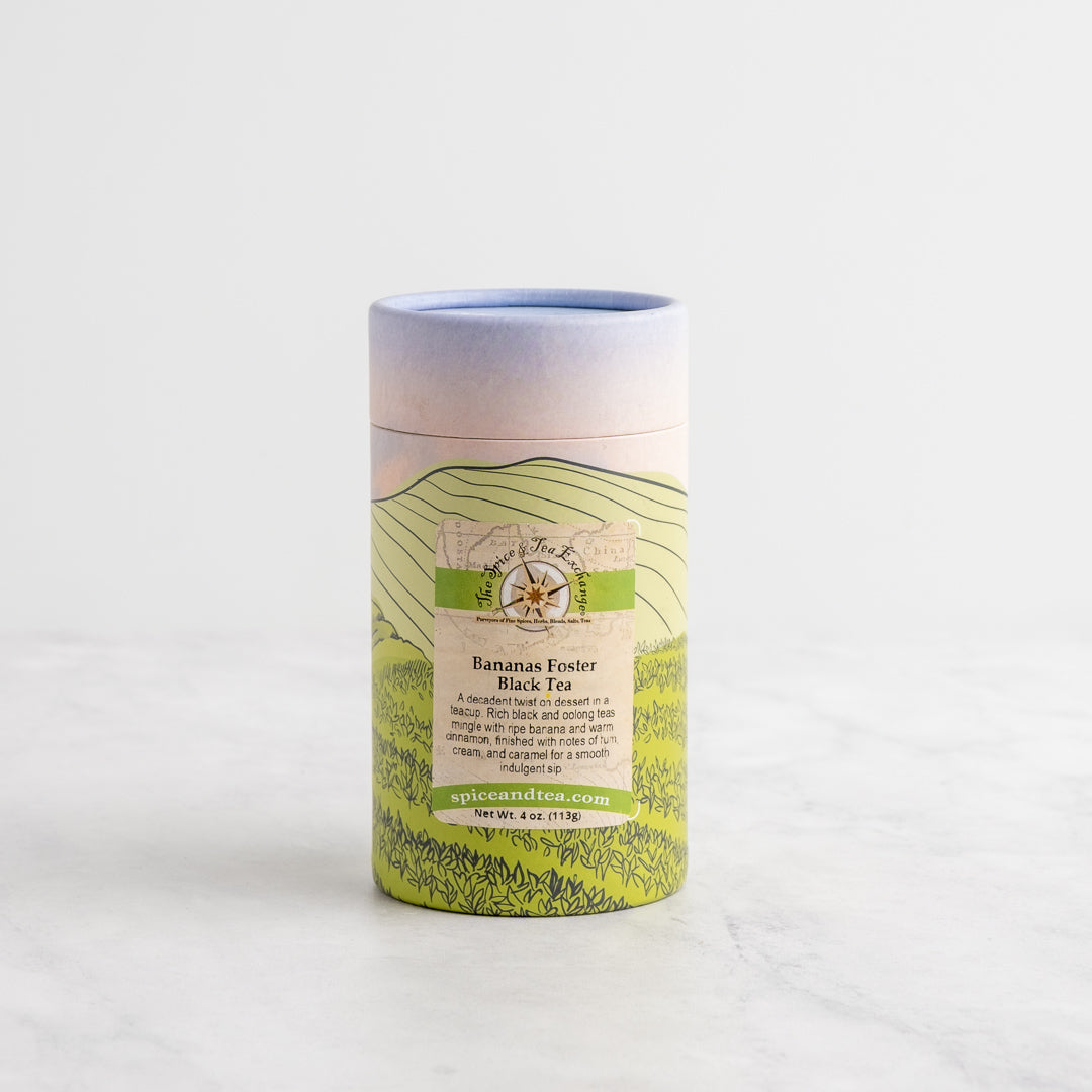 A cylindrical cardboard tube package of bananas foster black tea, featuring a pastel gradient top and a green illustrated label, set on a light marble surface against a white background.