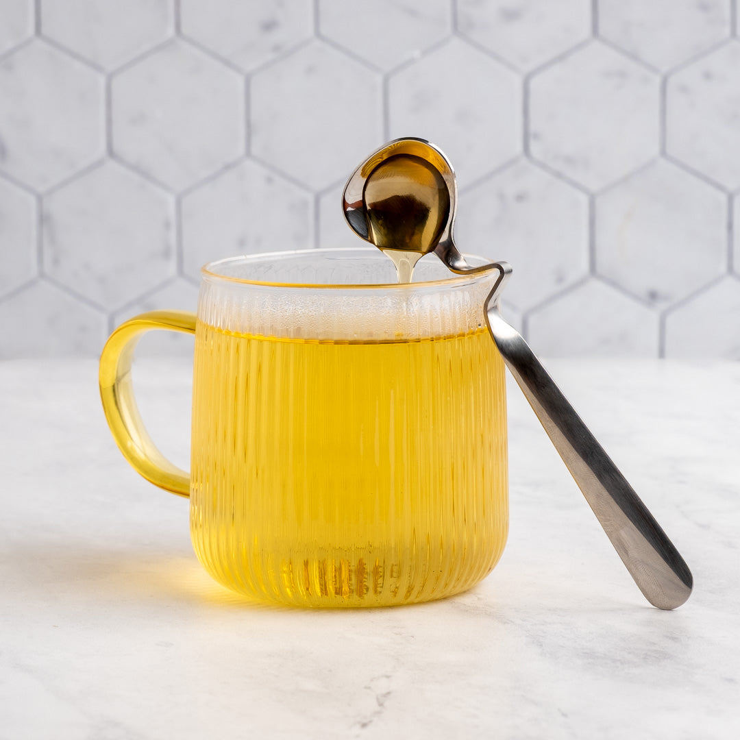 A ribbed glass mug with a translucent yellow handle and body, filled with tea, with a metal spoon resting on the rim and honey dripping into the mug, set against a white hexagonal tile background.