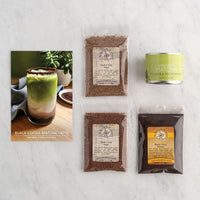 A flat-lay of a Black Cocoa Matcha Latte gift set on a white marble background, featuring a recipe card for a black cocoa matcha latte, two bags of dark cocoa powder, a tin of matcha tea extract, and a bag of black cocoa.