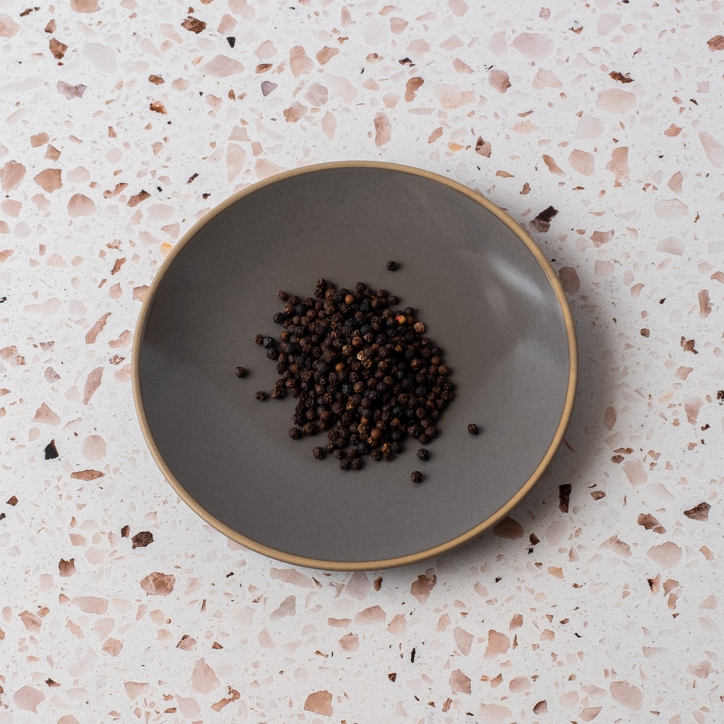 A grey ceramic plate featuring Smoked Black Peppercorn rests on a speckled, light terrazzo surface.